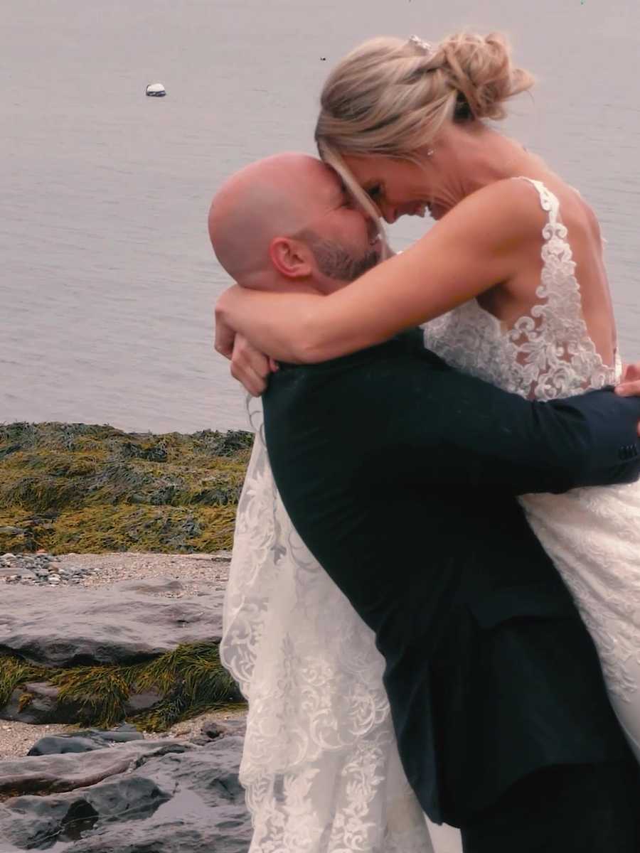Groom lifting the bride off her feet for a kiss on a rocky, seaweed-covered shoreline