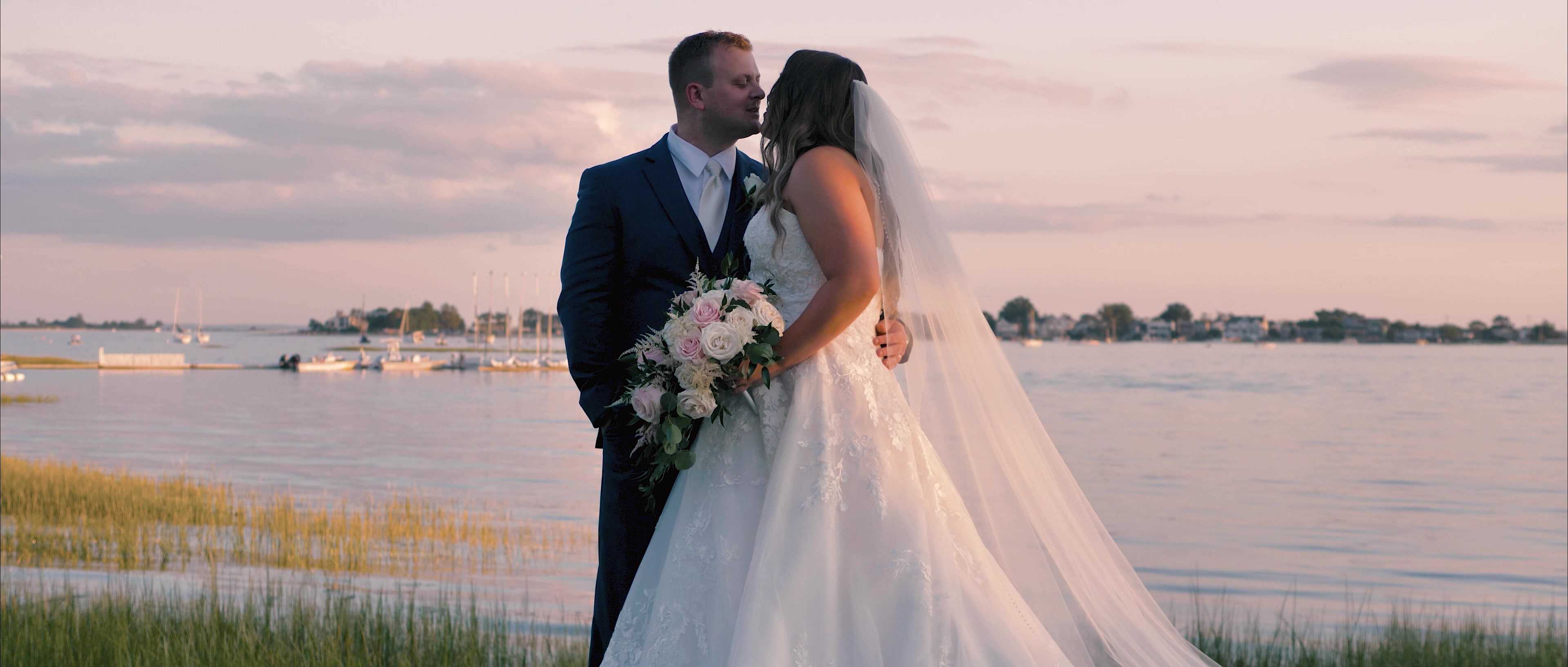 Couple with a bouquet at the water's edge under a pink sunset sky