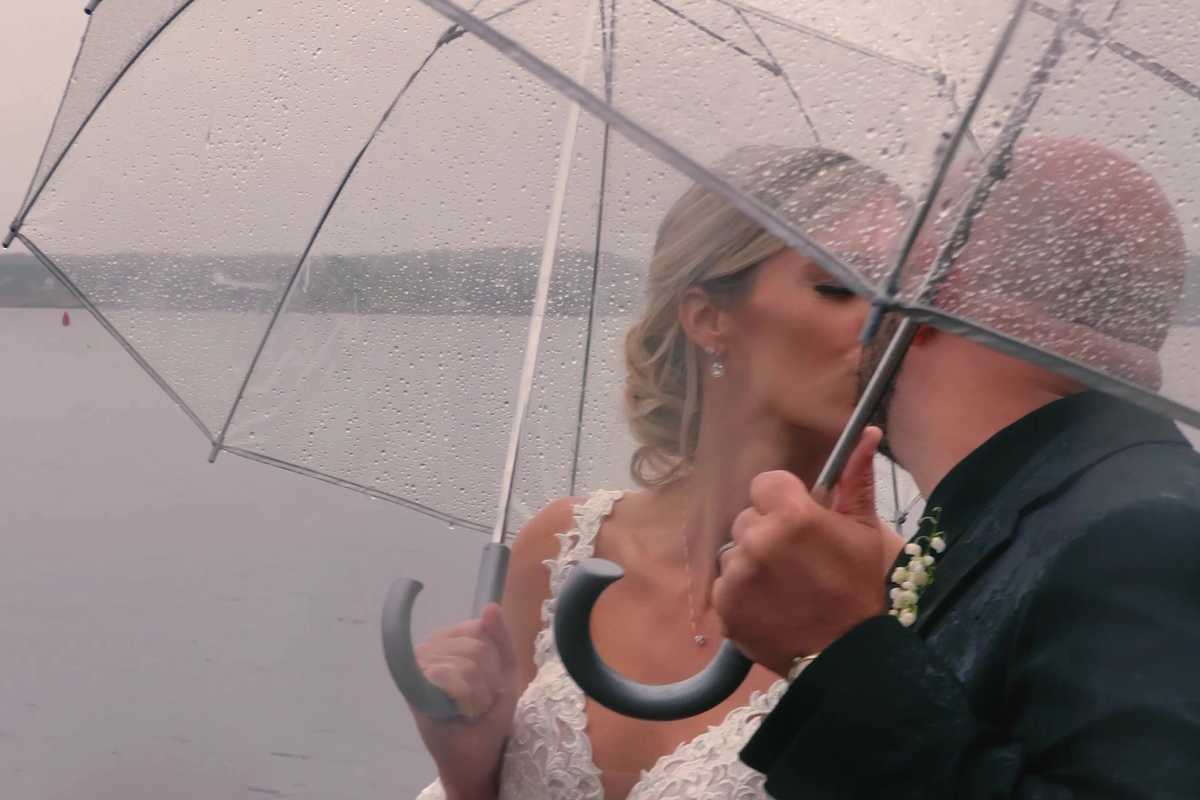Bride and groom kissing beneath a clear umbrella in the rain at the edge of a lake